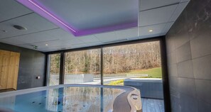 Un'area piscina moderna al coperto, con struttura in vetro, ampie finestre e vista su un paesaggio boschivo.