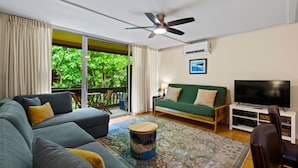 Sala de estar com sofá, televisão, ventilador de teto e vista para o verde lá fora.
