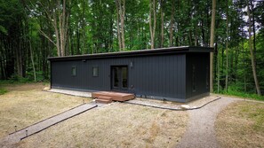 A small, dark-colored cabin with a door and windows, surrounded by a forest. - The Rae - a secluded & modern cabin (Mancelona)