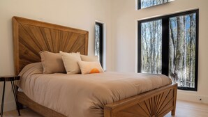Ein Bett mit einem Holz-Kopfteil, beige Bettwäsche und zwei Fenstern mit Blick auf einen Wald.