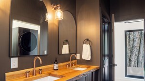 Ein Badezimmer mit zwei Waschbecken, einem großen Spiegel und Blick auf einen Wald durch ein Fenster.