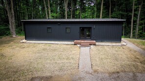 Eine moderne, dunkel gefärbte Holzhütte mit einer kleinen Veranda, umgeben von einem Wald.