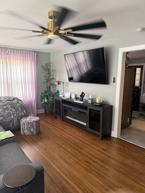 A modern living room with a ceiling fan, a flat-screen TV, a dark wood floor, a gray sofa, and a small round ottoman.
