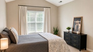 Une chambre à coucher avec un lit, une table de chevet, une commode, une fenêtre avec des rideaux et une plante.