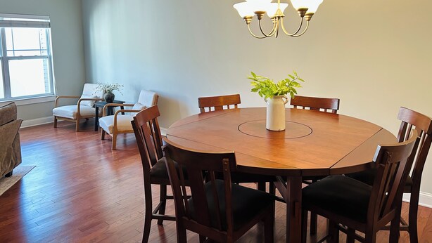Un coin repas comprenant une table ronde en bois, six chaises, un vase avec une plante et un lustre.