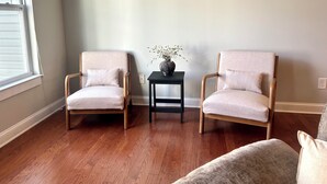 Une pièce avec deux fauteuils, une petite table sur laquelle est posé un vase, et une fenêtre avec des stores.
