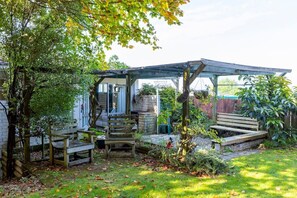 Een overdekt terras met houten banken en een tafel, omgeven door groen en een stenen muur.