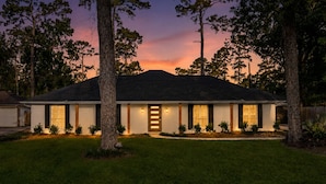 A two-story house with a dark roof, white exterior walls, and a front door with a glass panel. There are trees in the front yard and a clear sky in the background. - Spring Oasis: Heated Pool, Spa • Pickleball • Mini Golf • Basketball • Sleeps 12 (Spring)