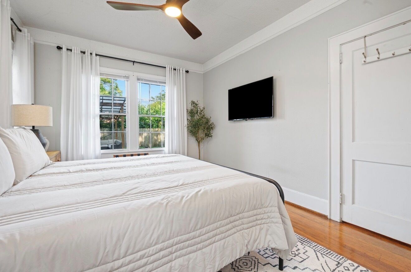 A bedroom with a bed, a ceiling fan, a TV, a window with curtains, and a door.