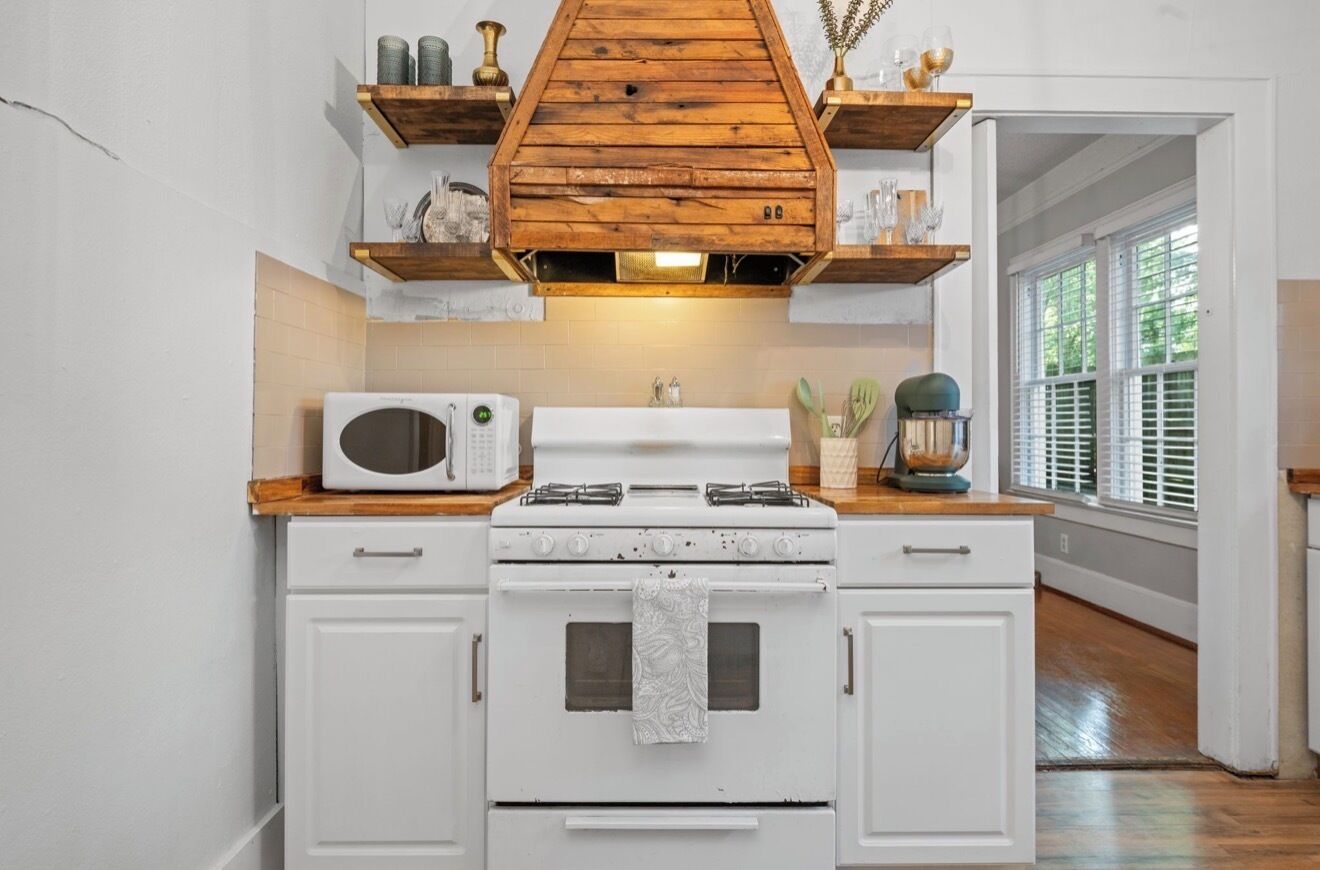 Une cuisine avec des meubles blancs, une cuisinière blanche, un four à micro-ondes, des étagères en bois et une fenêtre avec des stores.