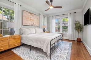 Quarto com cama, ventoinha no teto, televisão, janela com cortinas, abajur e uma planta.