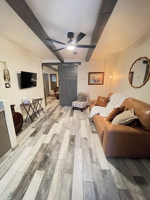 A living room with a brown leather couch, a patterned ottoman, a television mounted on the wall, and a ceiling fan. - Cindy's Cozy Casita (Green Valley)