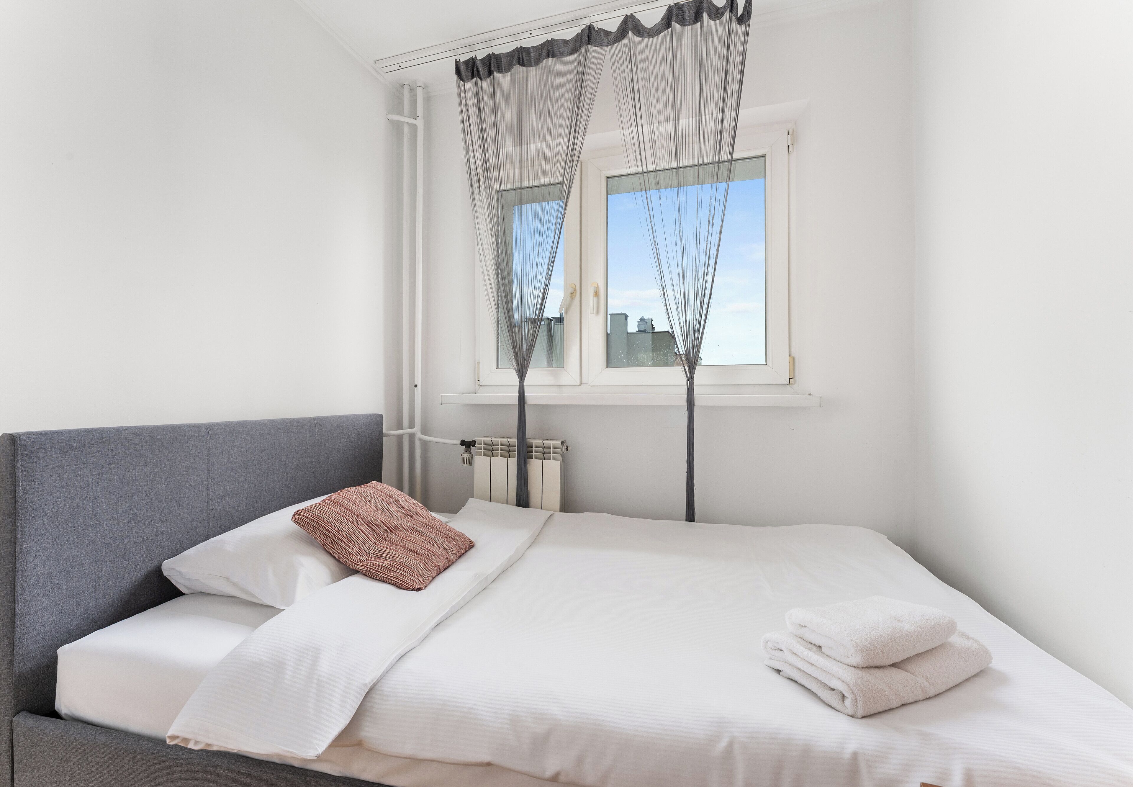 A bedroom with a bed, a grey headboard, a window with sheer curtains, and a radiator below the window.