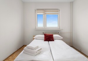 Una cama individual con ropa de cama blanca y una almohada roja, una ventana con persianas y un radiador sobre la cama.