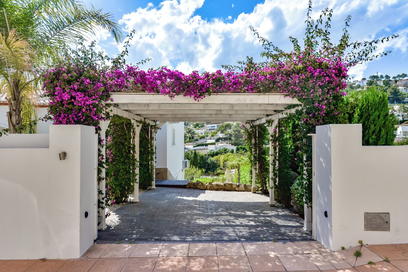 A covered walkway with a white structure, surrounded by greenery and flowering plants.