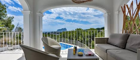 A balcony with a pool, a sofa, a chair, and a table with drinks and a book.