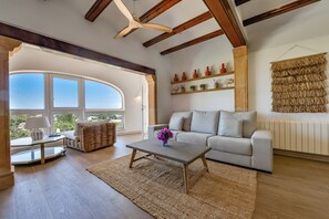 A spacious living room with a large window, a sofa, a coffee table, and a ceiling fan. - Miramar - By Almarina Villas (JÁVEA)