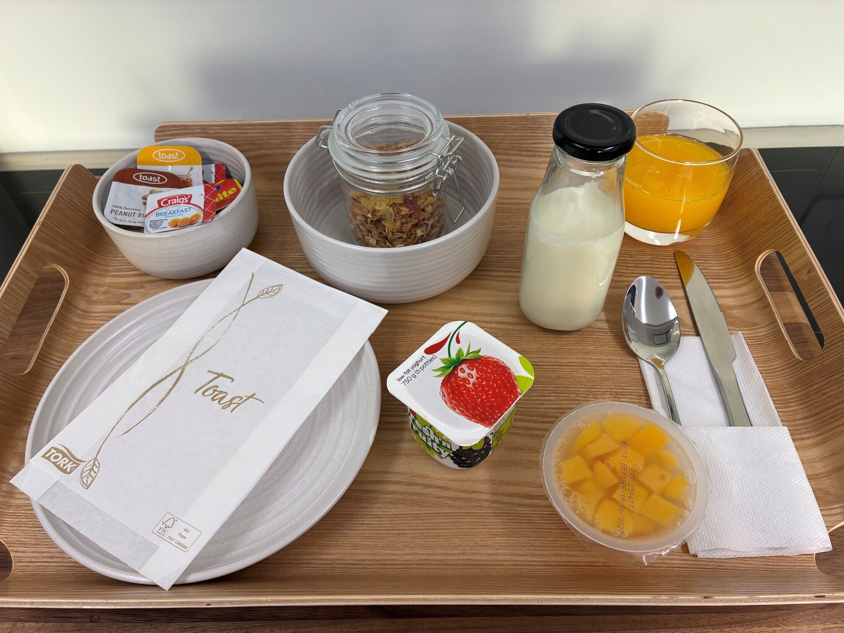 Ein Frühstücksbrett mit Toast, Müsli, Milch, Orangensaft, Joghurt und einer Obstschale.