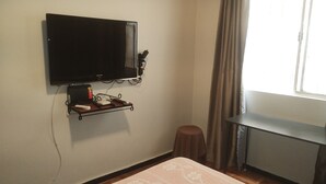 Una habitación con una televisión montada en la pared, una mesita con una bandeja, un taburete y una ventana con cortinas.