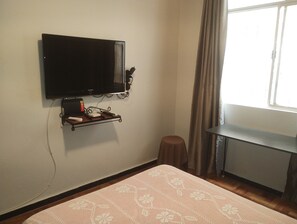 Ein Zimmer mit einem an der Wand befestigten Fernseher, einem kleinen Tisch mit Tablett, einem Hocker und einem Fenster mit Vorhängen.