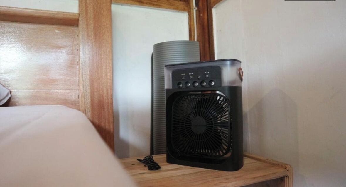 A bed with a wooden headboard, a small table with a black fan, and a cylindrical object in the background.