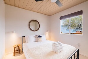 A bedroom with a bed, a round mirror, a window with a view, a small stool, and a bedside table. - Desert Oasis w/ Pool, Firepit~Stargazing! (Joshua Tree)