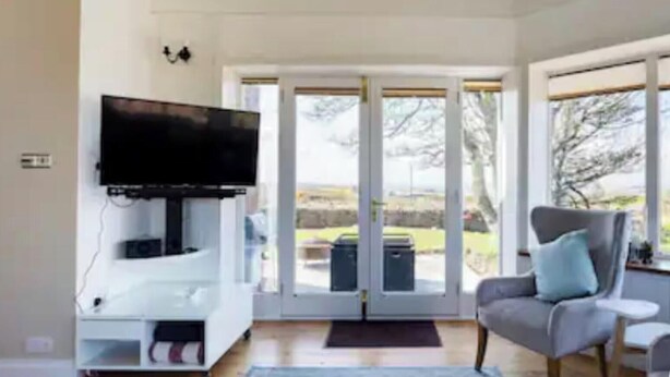 A modern living room with a flat-screen TV, a grey sofa, and a large window offering a view of the outdoors. - 4 Bed House - Sleeps 10 - Pets - Garden - Parking (Banff)
