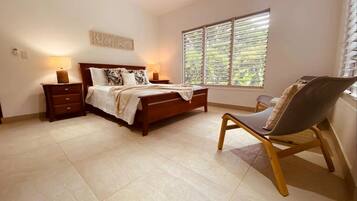 Une chambre à coucher comprenant un lit, deux tables de chevet, une chaise, une fenêtre avec des stores et une œuvre d’art accrochée au mur.