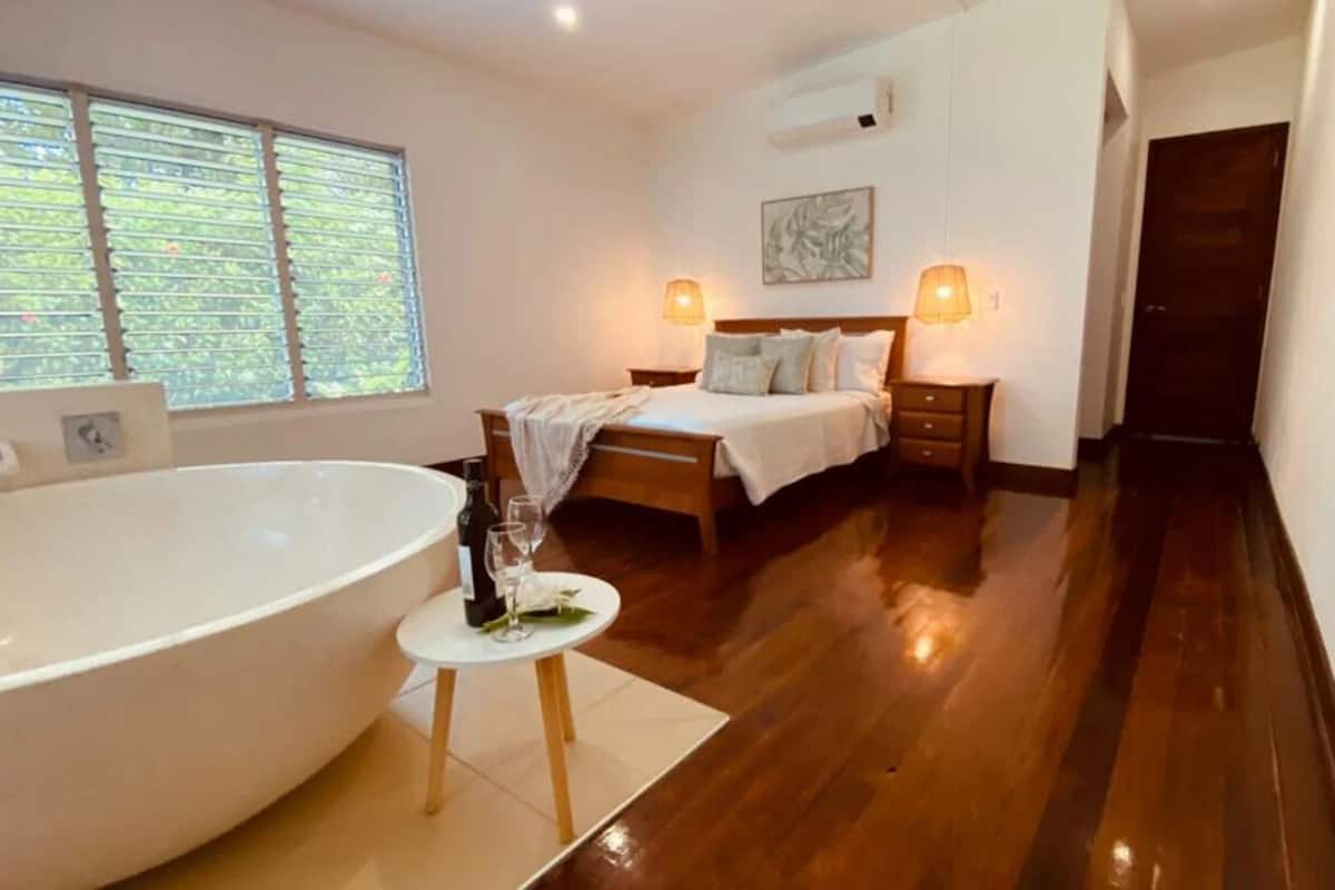 A bedroom with a large bathtub, a bed with a headboard, bedside lamps, and a window with blinds.