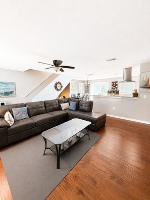 A modern living room with a sectional sofa, a glass coffee table, wooden flooring, a ceiling fan, and a kitchen area with a stove and window. - Brookside Oasis Leisure Bradenton Stay (Bradenton)