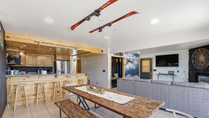 Eine moderne Küche mit Holzschränken, einer zentralen Kochinsel mit Hockern, einem Esstisch mit Bänken und einem Wohnbereich mit Sofa und Kamin.