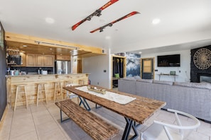 A modern kitchen with wooden cabinets, a central island with stools, a dining table with benches, and a living area with a sofa and fireplace.