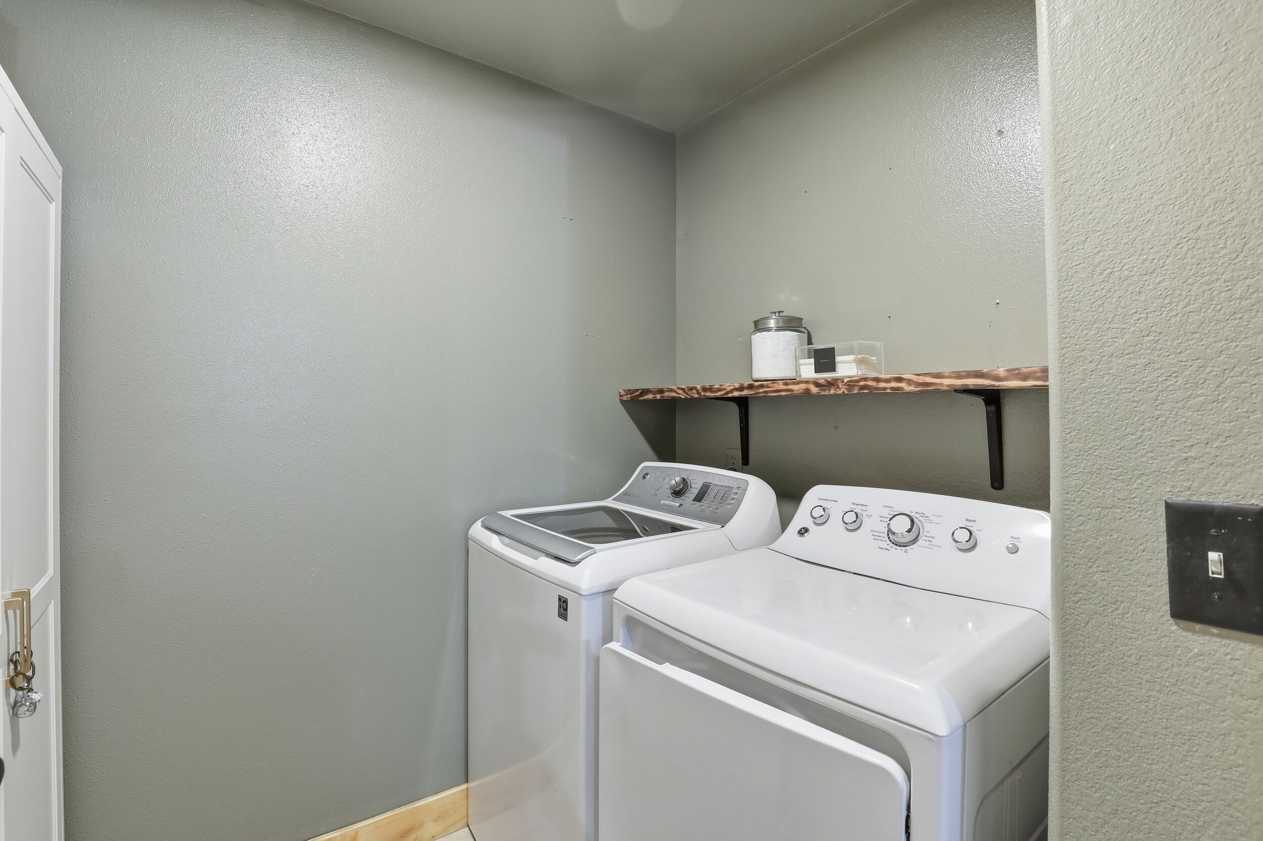 A laundry room with two white washing machines, a shelf with a container, and a door.
