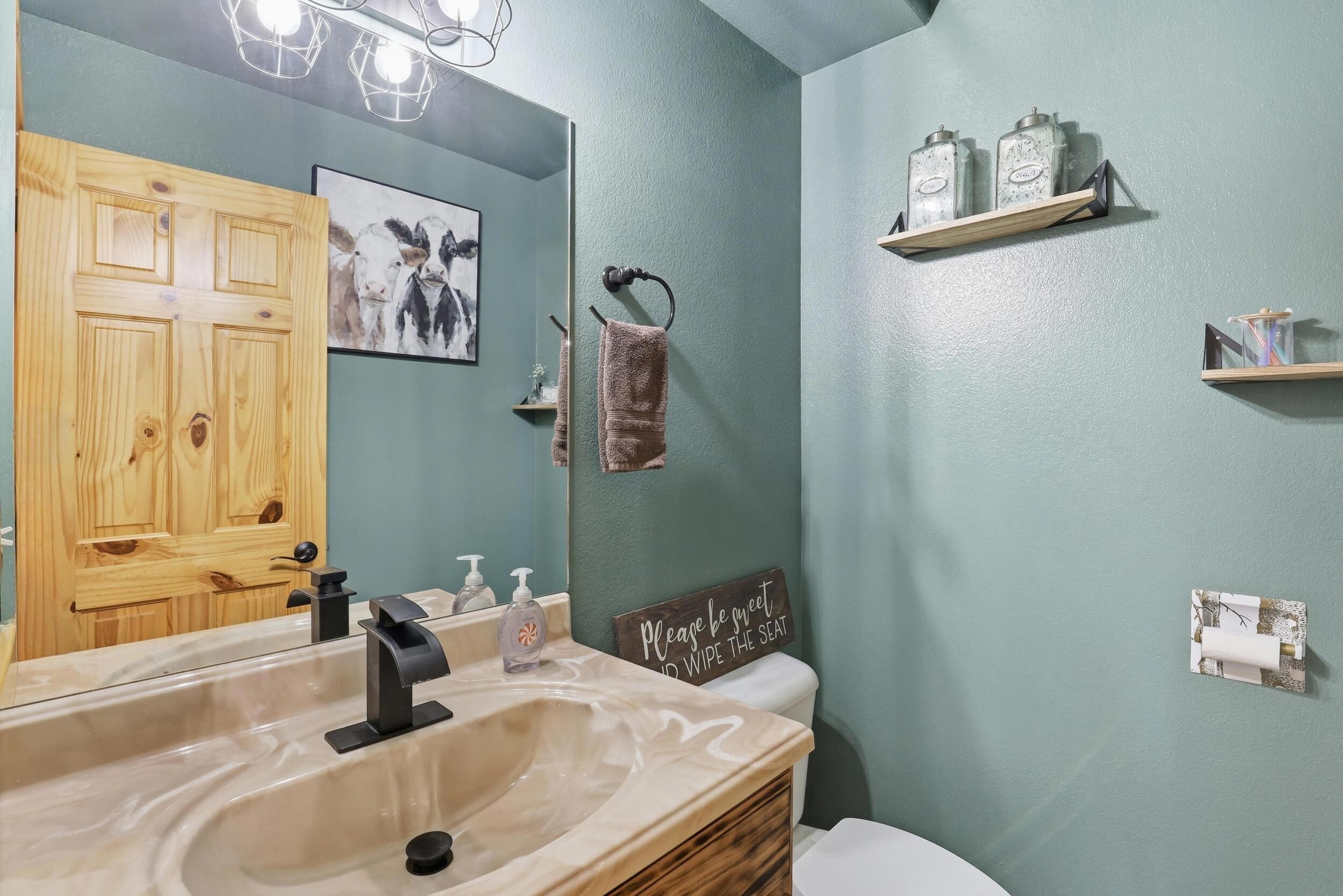 A bathroom with a wooden door, a sink with a mirror, a towel rack, and a sign on the wall.