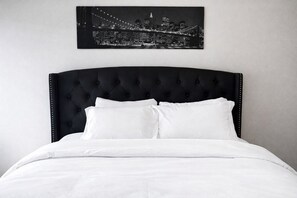 A bed with a tufted headboard, white bedding, and a cityscape photograph above.