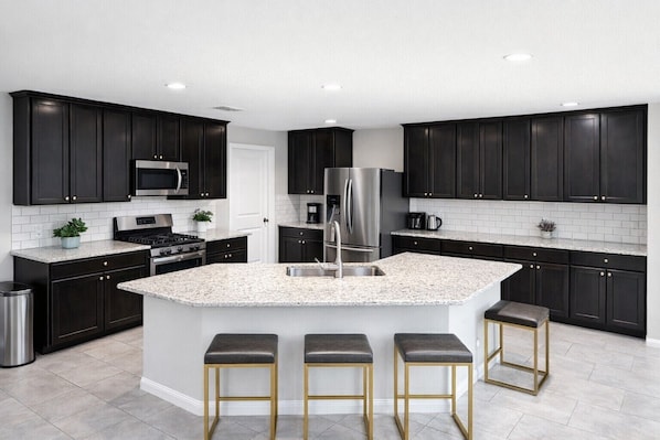 A modern kitchen with dark cabinetry, a central island with stools, stainless steel appliances, and a white tiled backsplash.