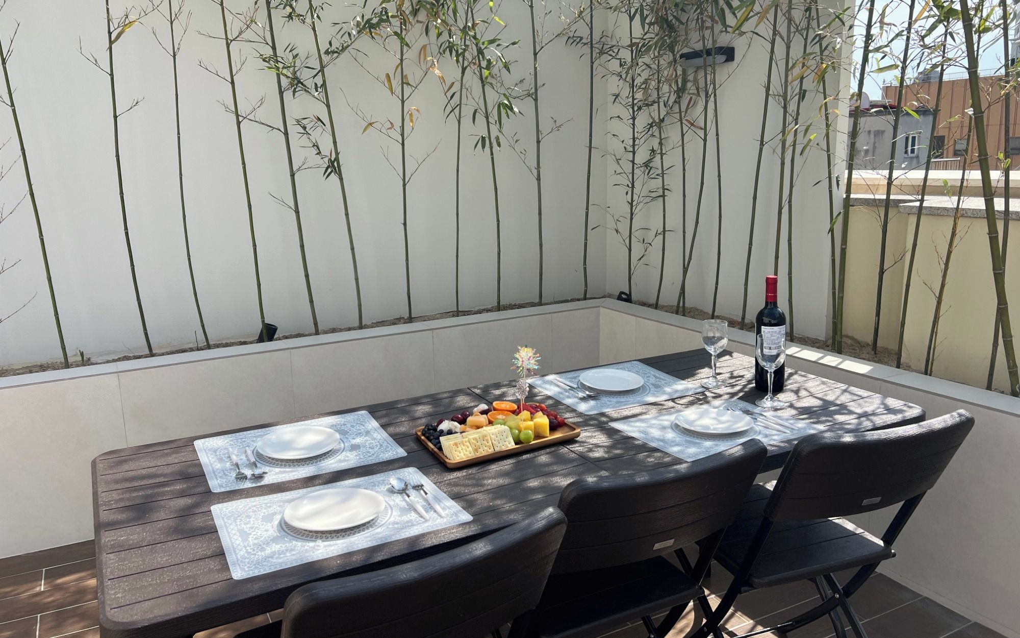 A dining table set outdoors with a bottle of wine, plates, cutlery, and a fruit platter.