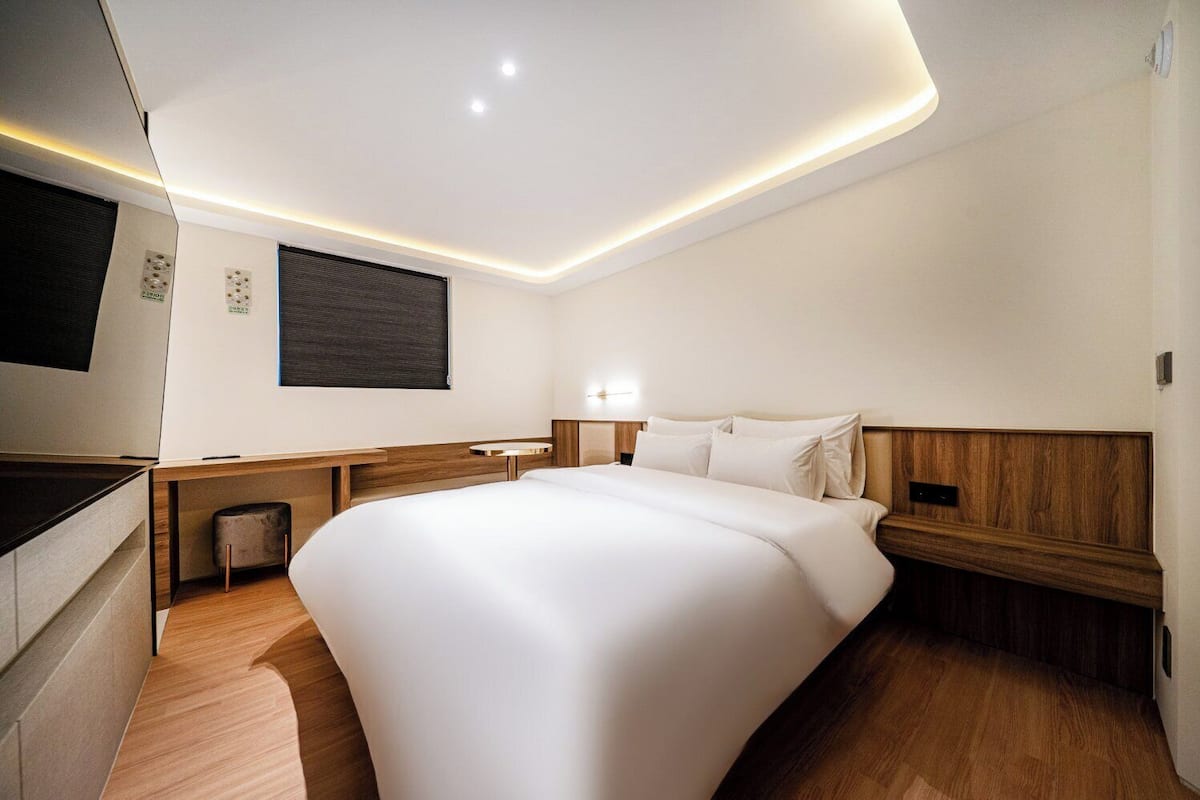 Una habitación de hotel moderna con una cama grande, cabecera de madera, mesitas de noche, televisor de pantalla plana y una lámpara fijada a la pared.