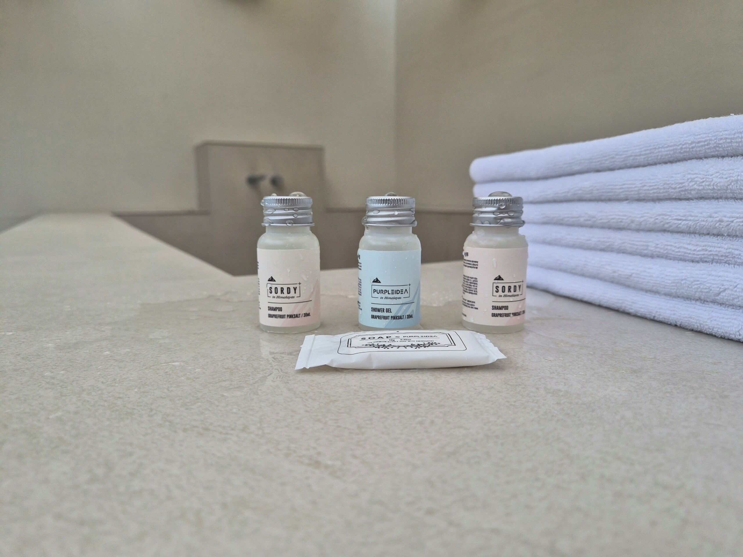 Three toiletry bottles on a bathroom counter, a folded towel, and a packet of soap.
