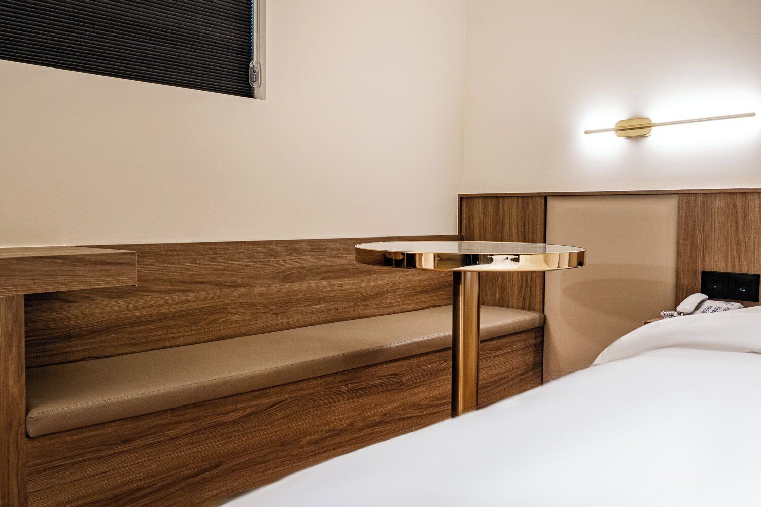 Een hotelkamer met een houten bankje, een klein rond tafeltje, een bed en een wandlamp.