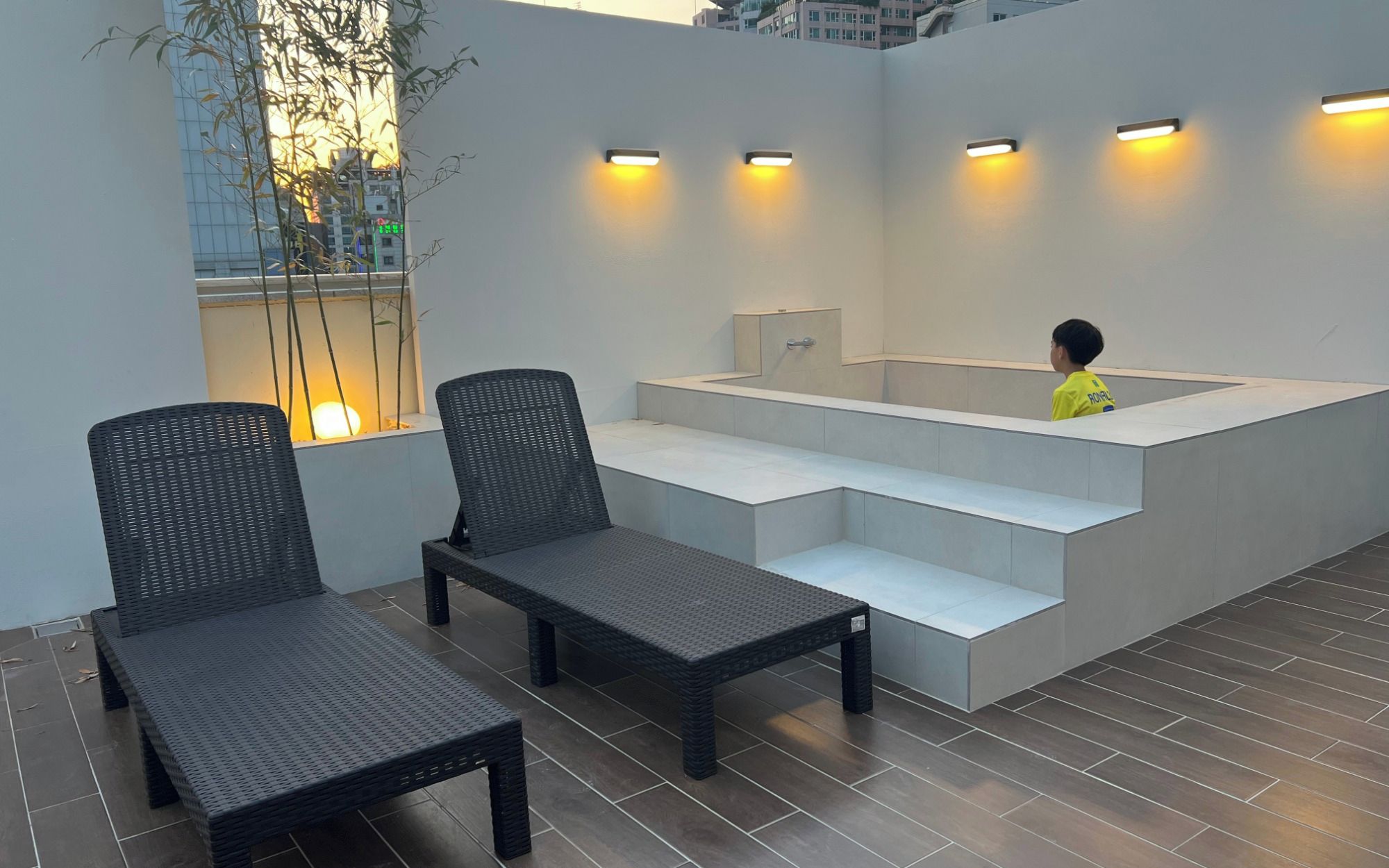 A modern outdoor seating area with two wicker chairs and a bench, a built-in bench with steps, and a wall-mounted light fixture.