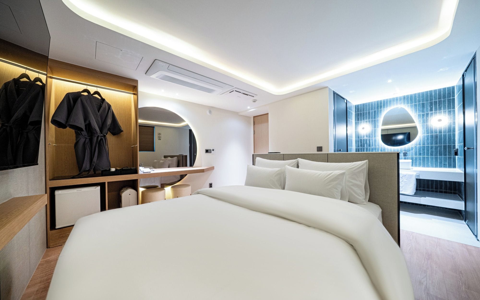 Une chambre d’hôtel moderne avec un grand lit, une coiffeuse avec un miroir et une salle de bain aux carreaux bleus.