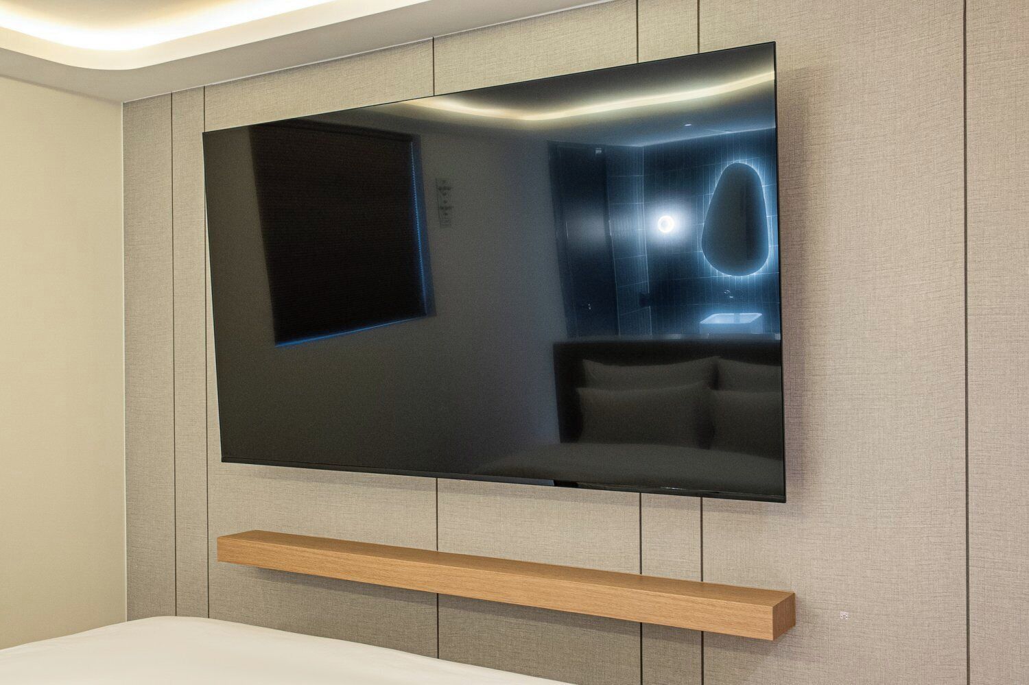 A modern room with a large flat-screen TV mounted on a textured wall, a wooden shelf below, and a dark-colored sofa.