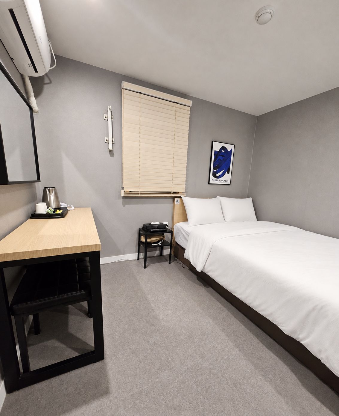 Quarto de hotel com uma cama de solteiro, escrivaninha, cadeira, ar condicionado, janela com persianas e uma obra de arte na parede.