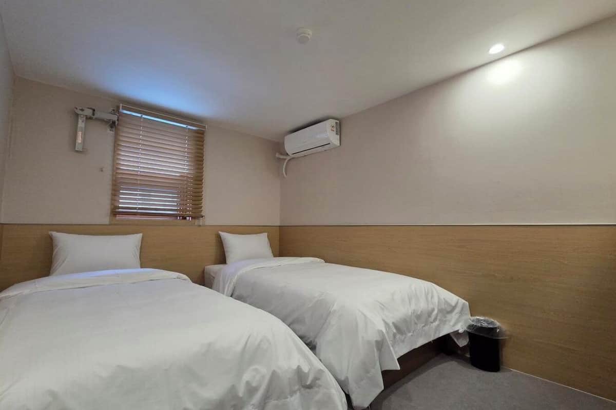 Une chambre d’hôtel avec deux lits, un climatiseur fixé au plafond, une fenêtre avec des stores et une petite poubelle.