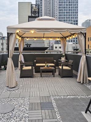 A rooftop terrace with a covered seating area, including a sofa and chairs, and a table with a bottle and snacks. - Cheonho T Hotel (Seoul)