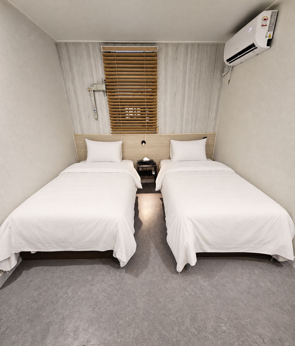 A hotel room with two single beds, a wooden headboard, a window with blinds, an air conditioning unit, and a grey carpet.
