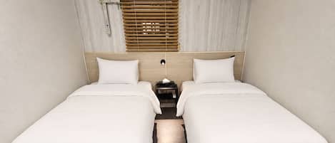 Habitación de hotel con dos camas individuales, cabecera de madera, ventana con persianas, equipo de aire acondicionado y alfombra gris.