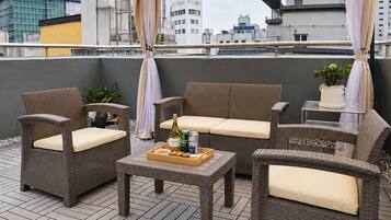 Eine Terrasse im Freien mit Korbmöbeln, einem Tisch mit einer Auswahl an Snacks und Getränken und Blick auf Stadtgebäude.