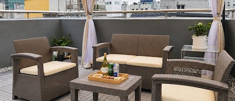 Un patio al aire libre con mobiliario de mimbre, una mesa con una bandeja de bocadillos y bebidas, y vistas a edificios de la ciudad.